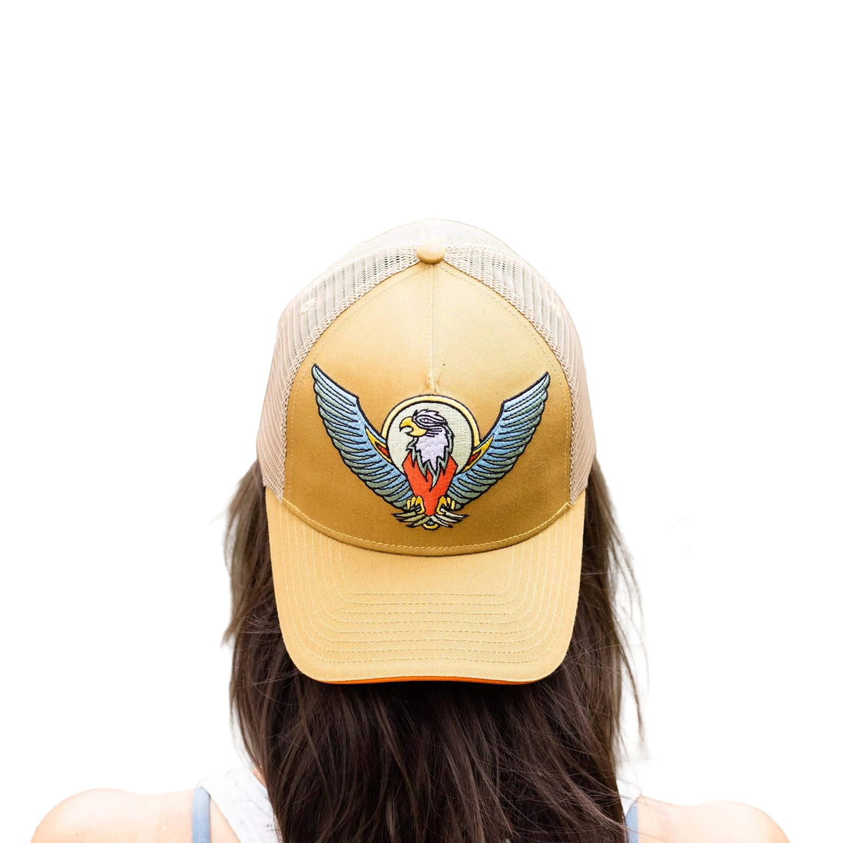 A person viewed from behind wearing a tan and beige trucker hat with an embroidered bald eagle logo, with a blurred outdoor background of water and greenery.