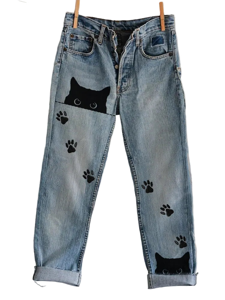 Blue jeans with black cat and paw print designs hanging on a line against a plain background