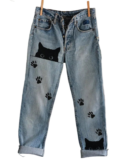 Blue jeans with black cat and paw print designs hanging on a line against a plain background