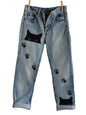 Blue jeans with black cat and paw print designs hanging on a line against a plain background