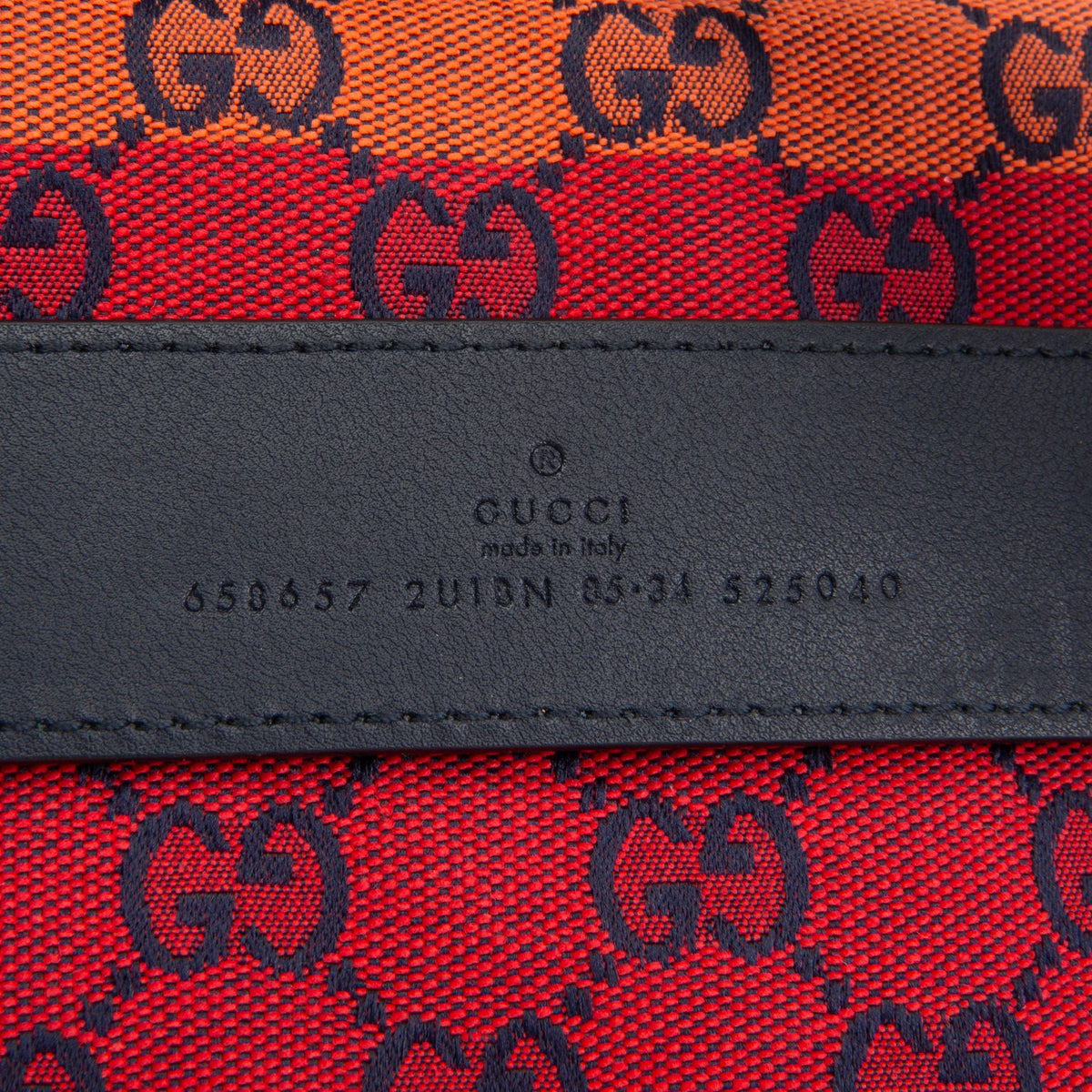 Close-up of a Gucci textile product showing the red and orange GG monogram canvas with a black leather band embossed with 'GUCCI made in Italy' and a UDIAN number.