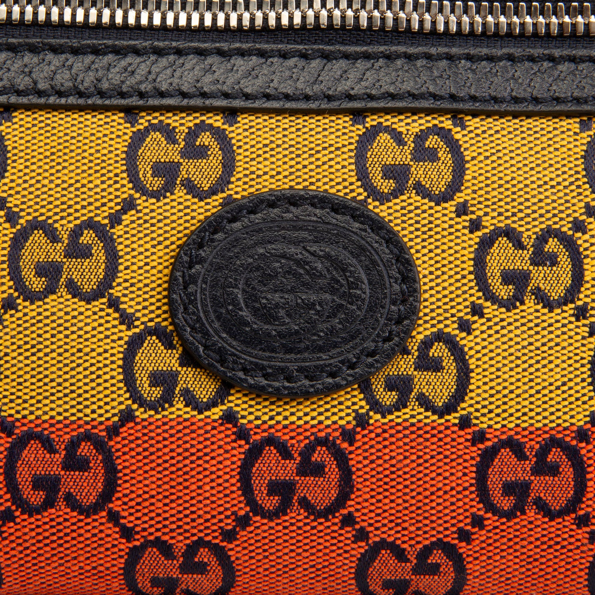 Close-up of a Gucci zip pouch featuring a yellow and orange canvas body with a black GG monogram pattern, a black leather zipper, and a central black oval GG logo patch.