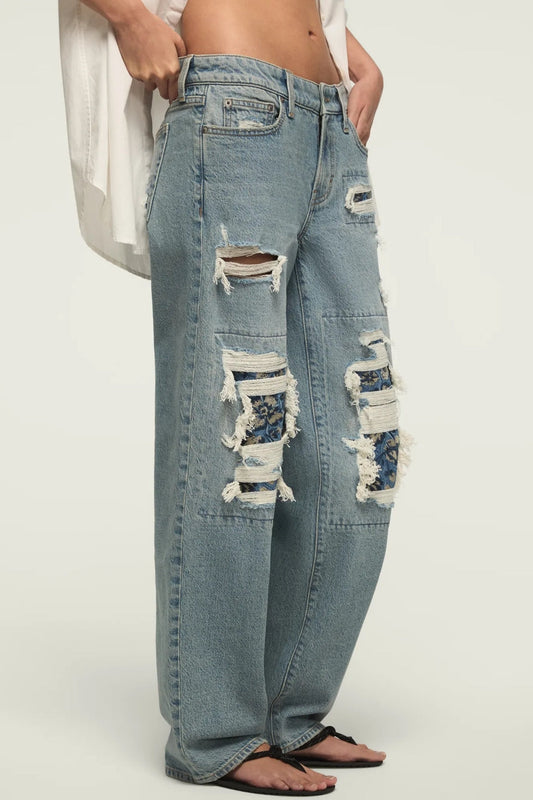 A person wearing light-wash, wide-leg distressed jeans with large ripped patches showing floral lining, paired with black flip-flops and a white top against a plain light background.