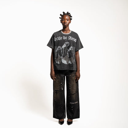 A model stands against a plain white studio background wearing a faded black graphic T-shirt with 'Style de Vita' text and a distressed black wide-leg pant with paint splatter details.