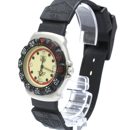 A Tag Heuer wristwatch with a black rubber strap, black bezel, and a cream-colored dial with red accents, displayed on a clear plastic stand against a white background.