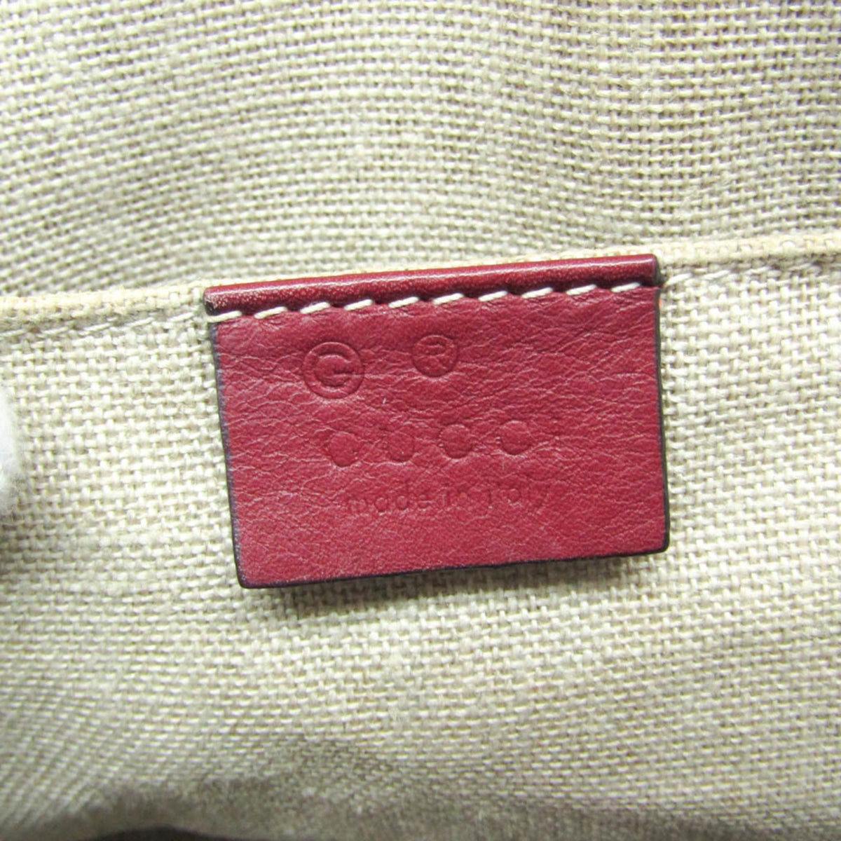 Close-up of a Gucci leather brand patch in dark red, stitched onto a beige woven canvas fabric.