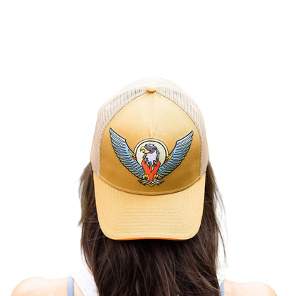 A person viewed from behind wearing a tan and beige trucker hat with an embroidered bald eagle logo, with a blurred outdoor background of water and greenery.
