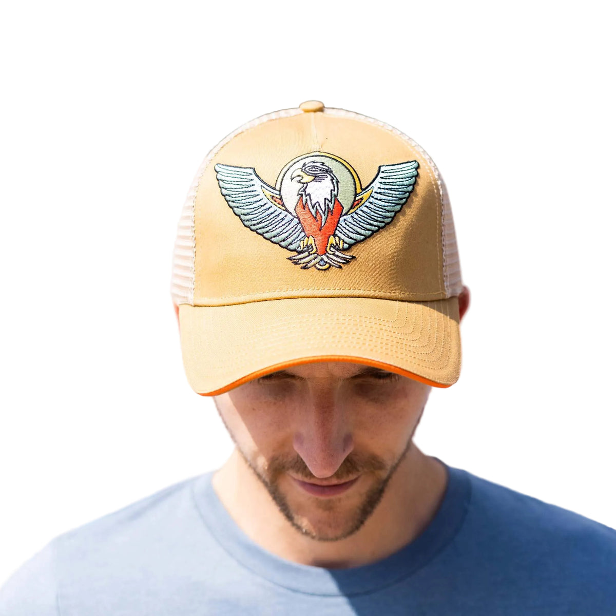A man wearing a tan trucker hat with a detailed eagle patch on the front, set against a colorful geometric background.
