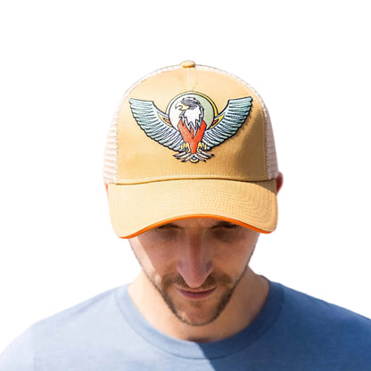 A man wearing a tan trucker hat with a detailed eagle patch on the front, set against a colorful geometric background.