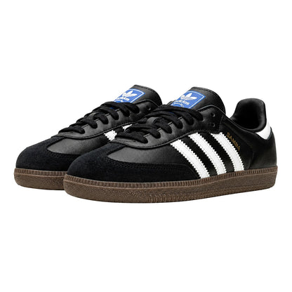 A pair of Adidas Samba sneakers in black suede and leather with white three-stripes, a blue tongue patch, and a brown gum sole, shown on a plain white background.
