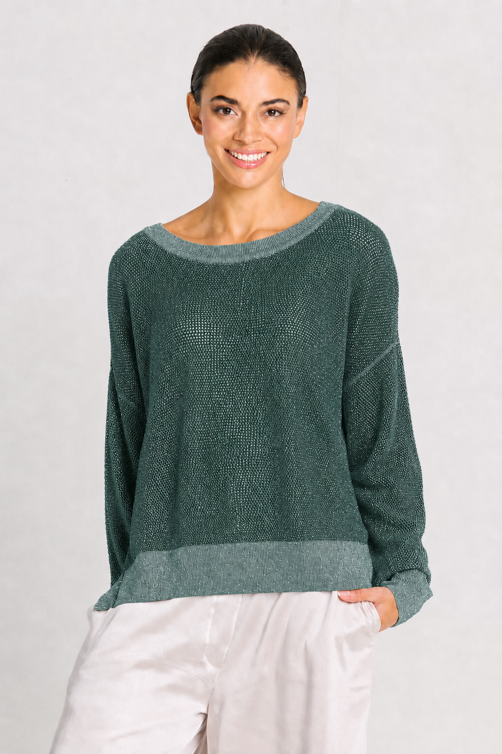A woman wearing a dark green knit sweater with a wide boat neckline and contrasting green ribbed trim at the collar and hem, paired with light-colored pants, against a light gray background.