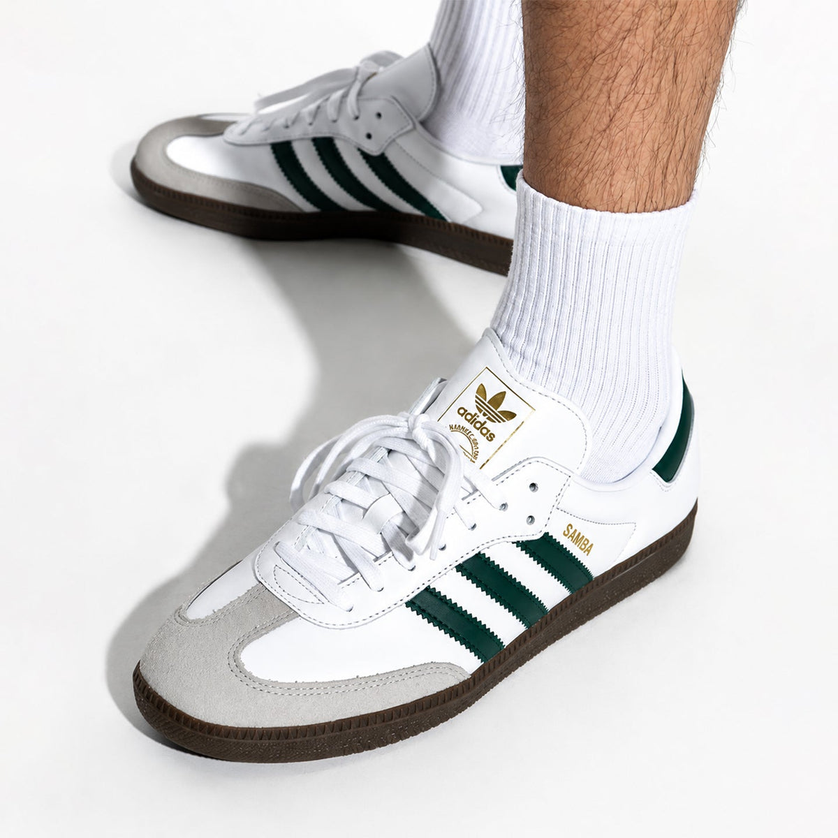 White Adidas Samba sneakers with dark green stripes, a light gray suede toe box, and a brown gum sole, shown on a model wearing white socks against a white background.
