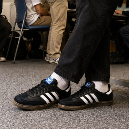A person wearing black Adidas Samba sneakers with white stripes, white socks, and black jeans, standing on a gray carpeted floor in a room with other people blurred in the background.