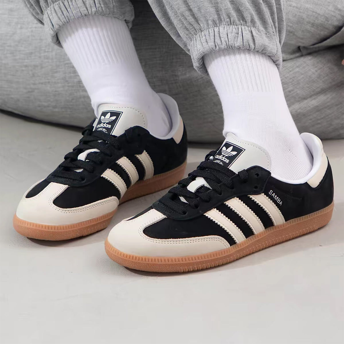 A pair of Adidas Samba sneakers in black and off-white with gum soles, worn with white crew socks and gray sweatpants.