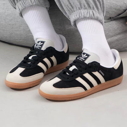 A pair of Adidas Samba sneakers in black and off-white with gum soles, worn with white crew socks and gray sweatpants.