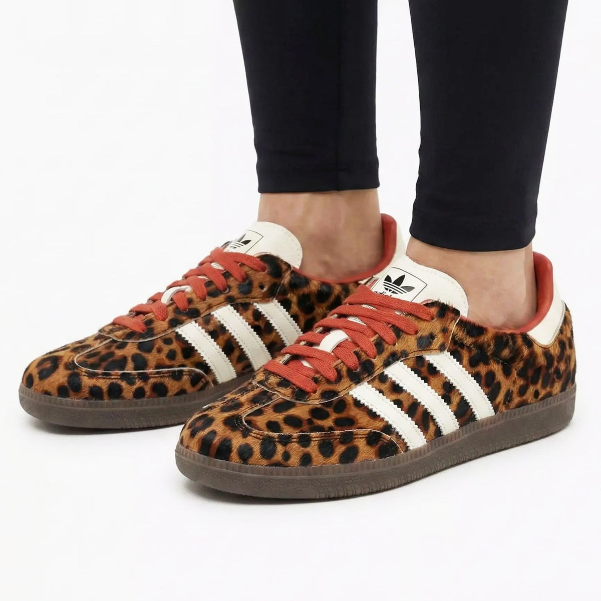 A pair of Adidas Gazelle sneakers with a leopard print pattern, white three-stripes, and orange laces, shown on a model wearing black pants against a white background.