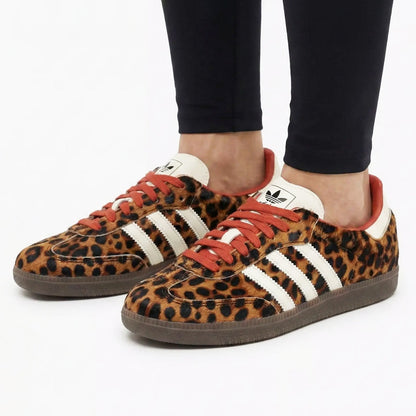 A pair of Adidas Gazelle sneakers with a leopard print pattern, white three-stripes, and orange laces, shown on a model wearing black pants against a white background.