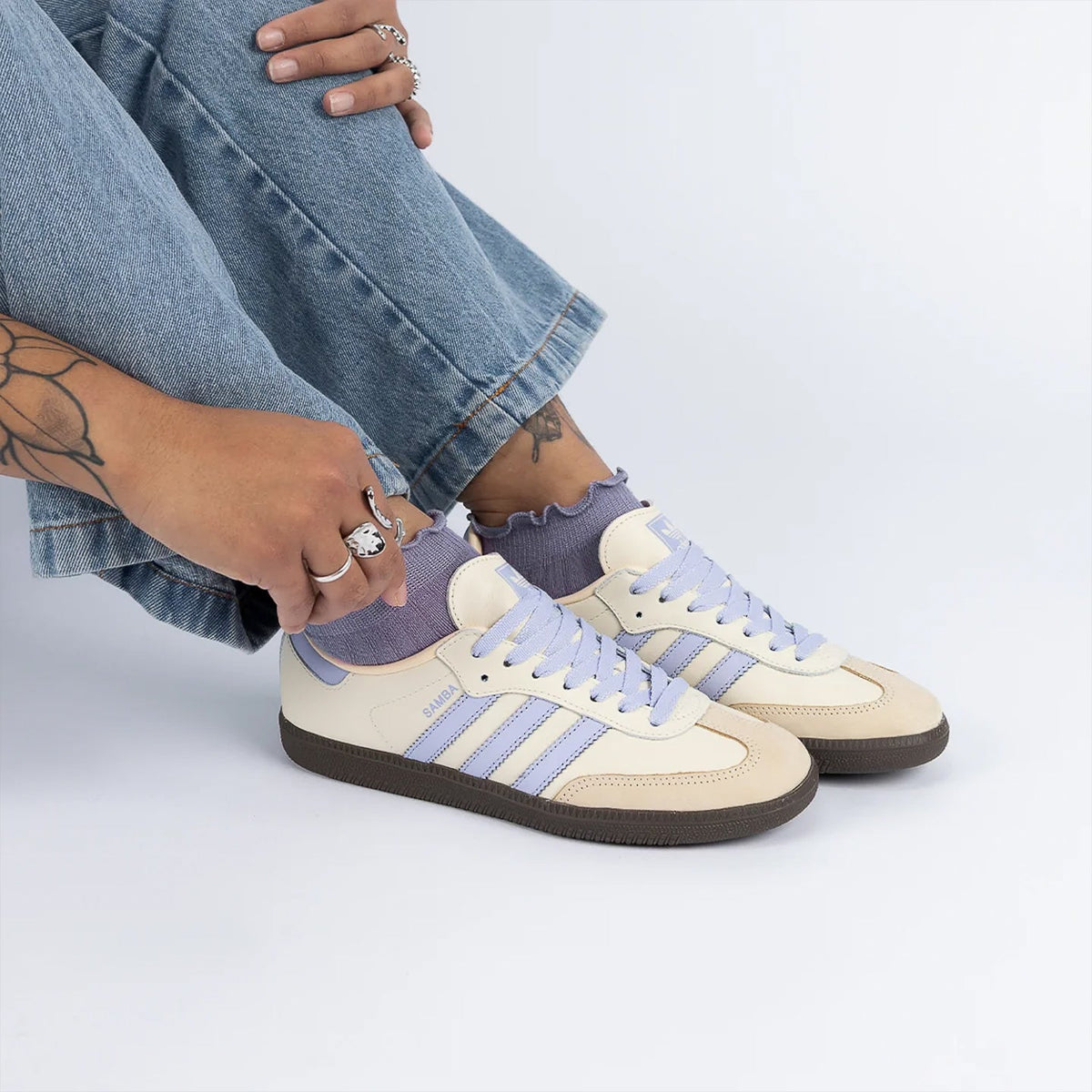 A person wearing light blue jeans and lavender ruffled socks is shown from the knees down, wearing a pair of Adidas Samba sneakers in cream and lavender. The person is wearing silver rings on their fingers and ankles.