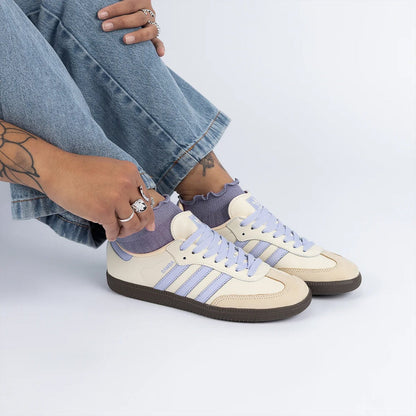 A person wearing light blue jeans and lavender ruffled socks is shown from the knees down, wearing a pair of Adidas Samba sneakers in cream and lavender. The person is wearing silver rings on their fingers and ankles.