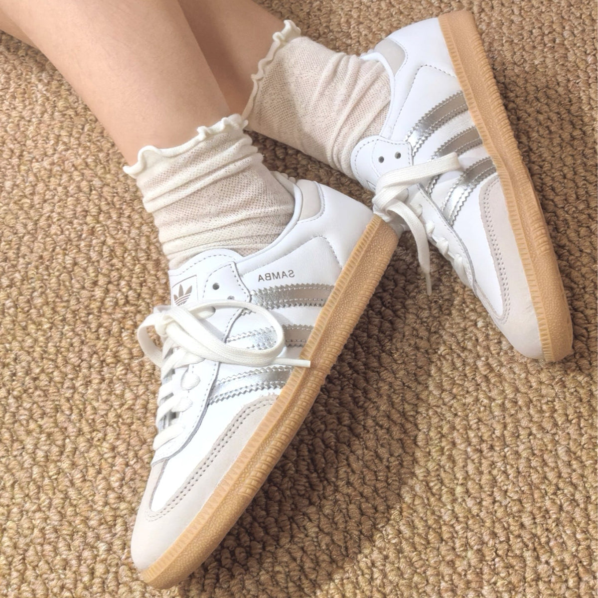 White Adidas Samba sneakers with silver stripes and beige gum soles, shown on a person's feet with cream ruffled socks on a textured beige carpet.