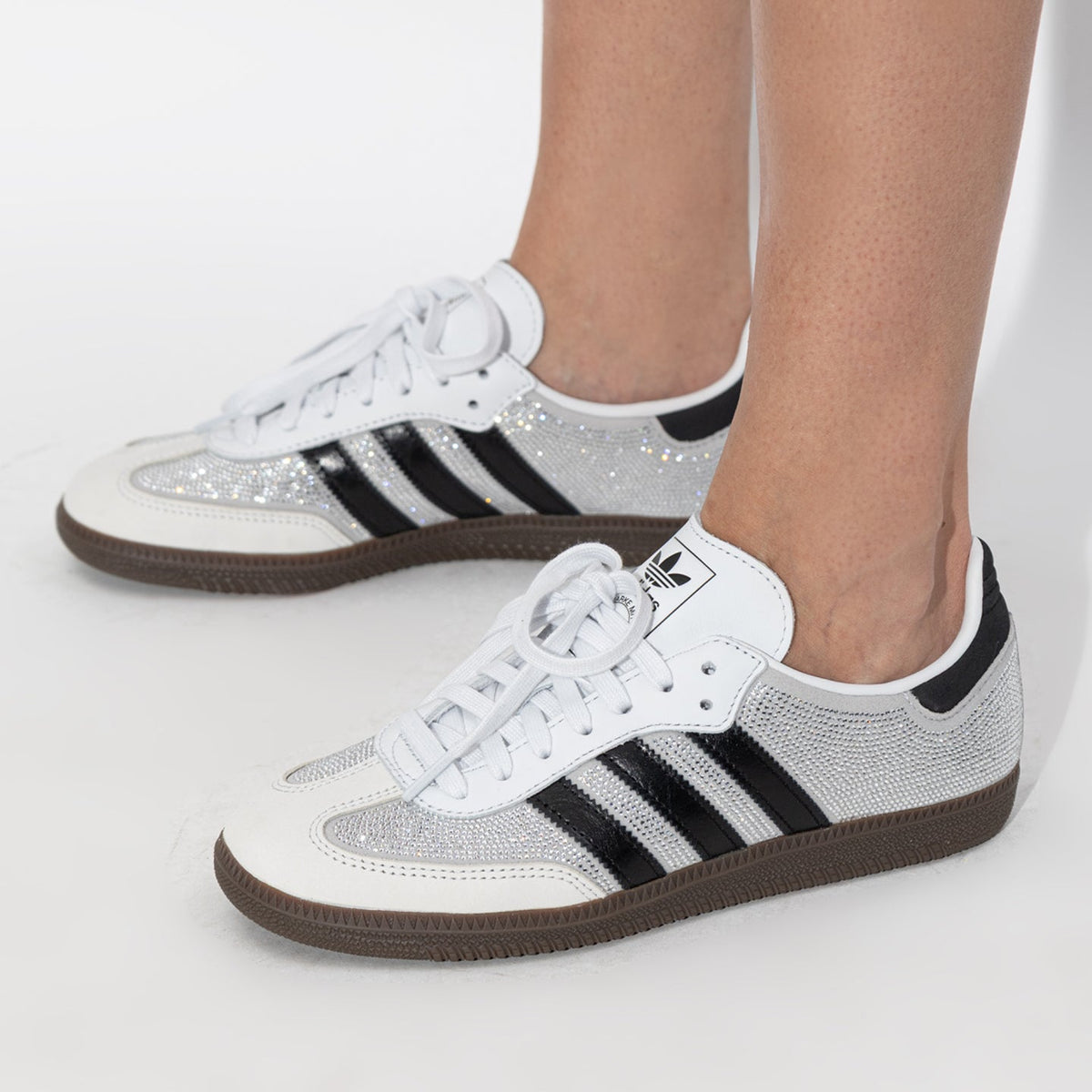 A pair of Adidas Samba sneakers in white and silver glitter with black stripes, shown on a model's feet against a white studio background.