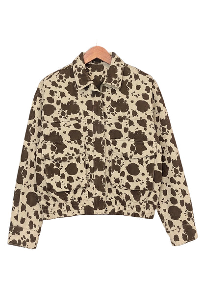 A cropped long-sleeve collared jacket with an all-over cow-print pattern in dark brown and beige, hanging on a wooden hanger against a rustic dark wood plank wall.