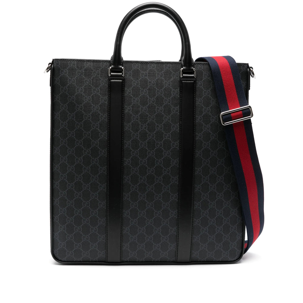 A black Gucci tote bag featuring the brand's signature GG monogram pattern, black leather trim, top handle, and a red and navy blue webbed shoulder strap with a silver metal buckle.