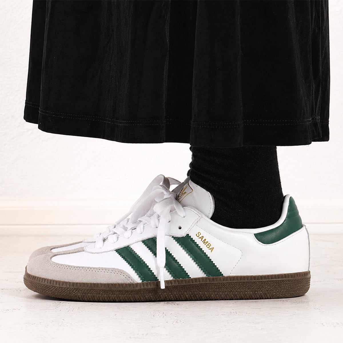 A white Adidas Samba sneaker with green stripes, a green heel tab, and gold 'SAMBA' text, shown on a model's foot with a black sock and black skirt visible.