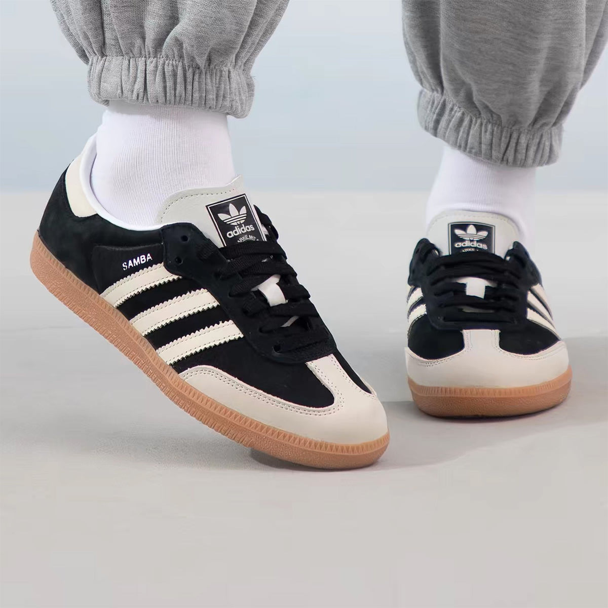 A pair of Adidas Samba sneakers in black, cream, and gum brown, shown being worn with white socks and gray sweatpants.