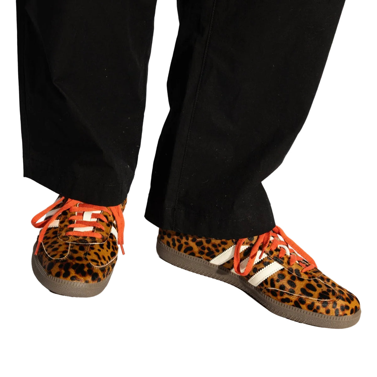 A person wearing black pants and leopard-print sneakers with bright orange laces and white side stripes, photographed against a clean white studio background.