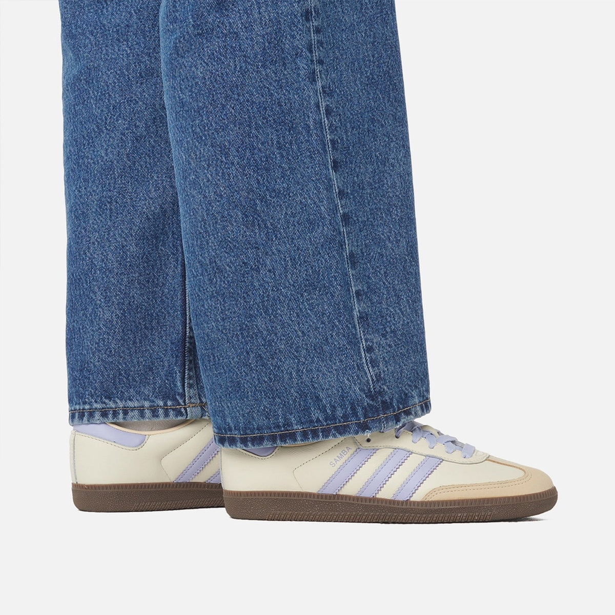 A close-up of a person's legs wearing blue jeans and cream-colored Adidas Samba sneakers with light purple stripes and brown soles, set against a plain light gray background.