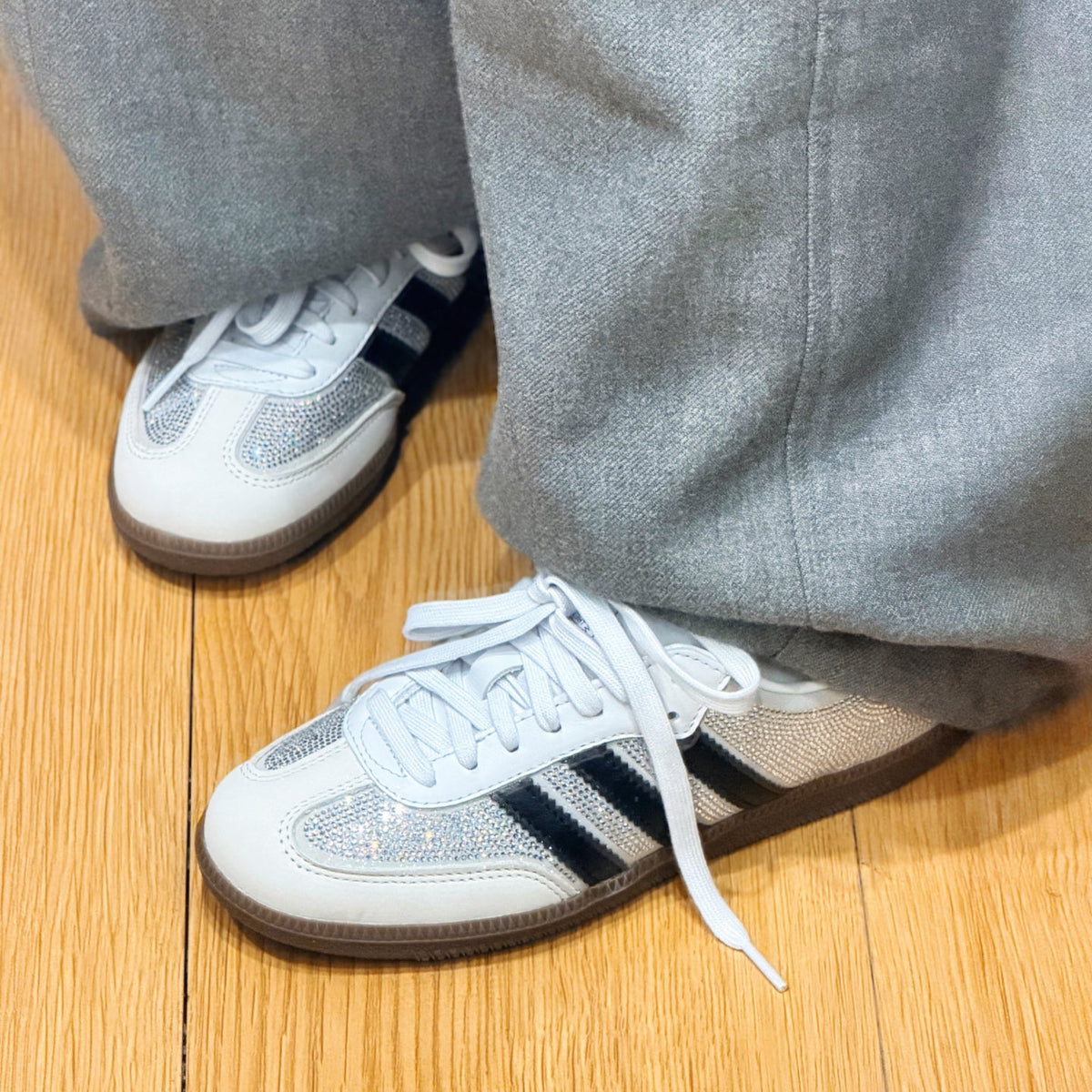 A pair of white Adidas Samba sneakers with silver glitter panels and black three-stripes, worn with gray sweatpants on a light wood floor.
