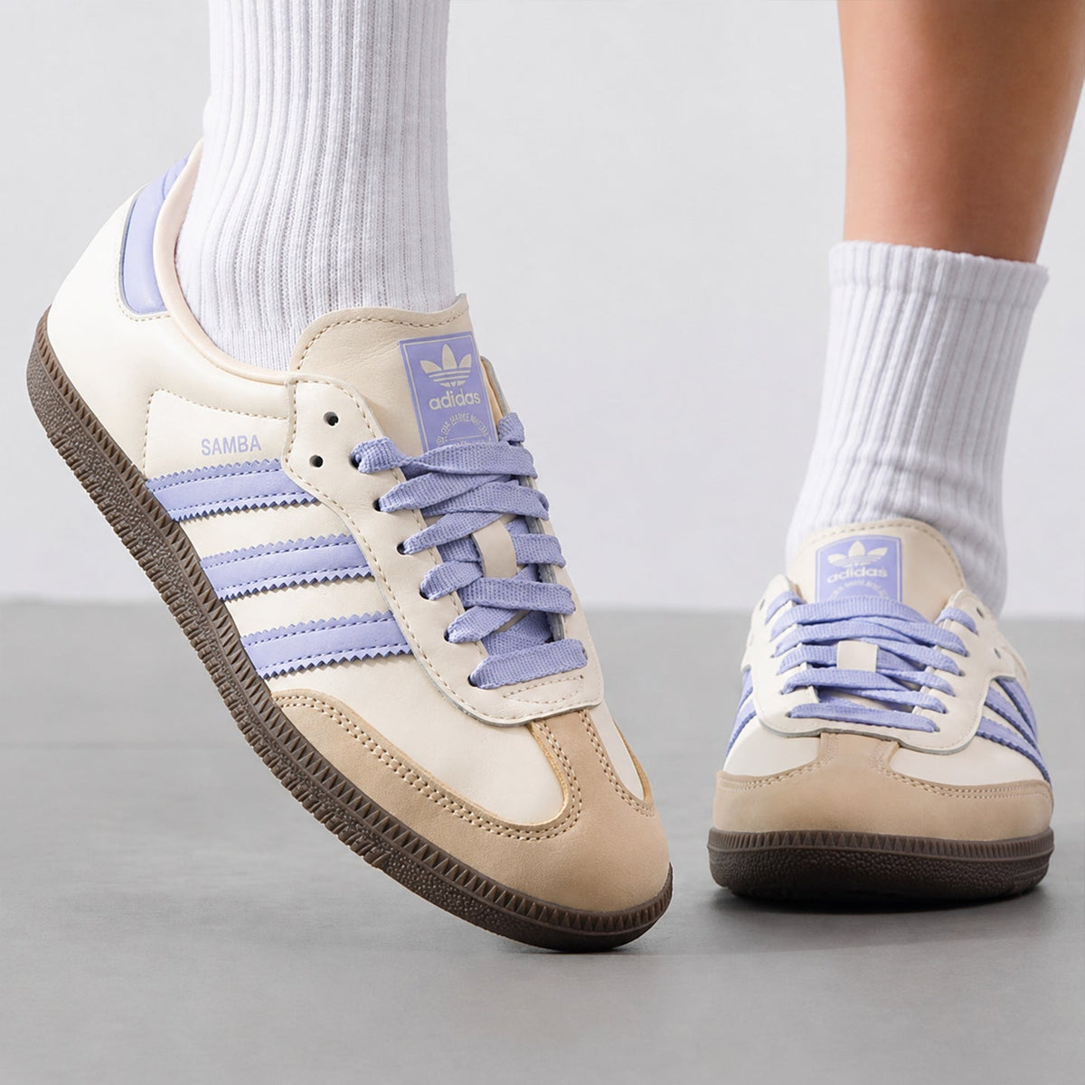 A pair of Adidas Samba sneakers in cream and light purple, shown on a person wearing white socks against a neutral gray studio background.