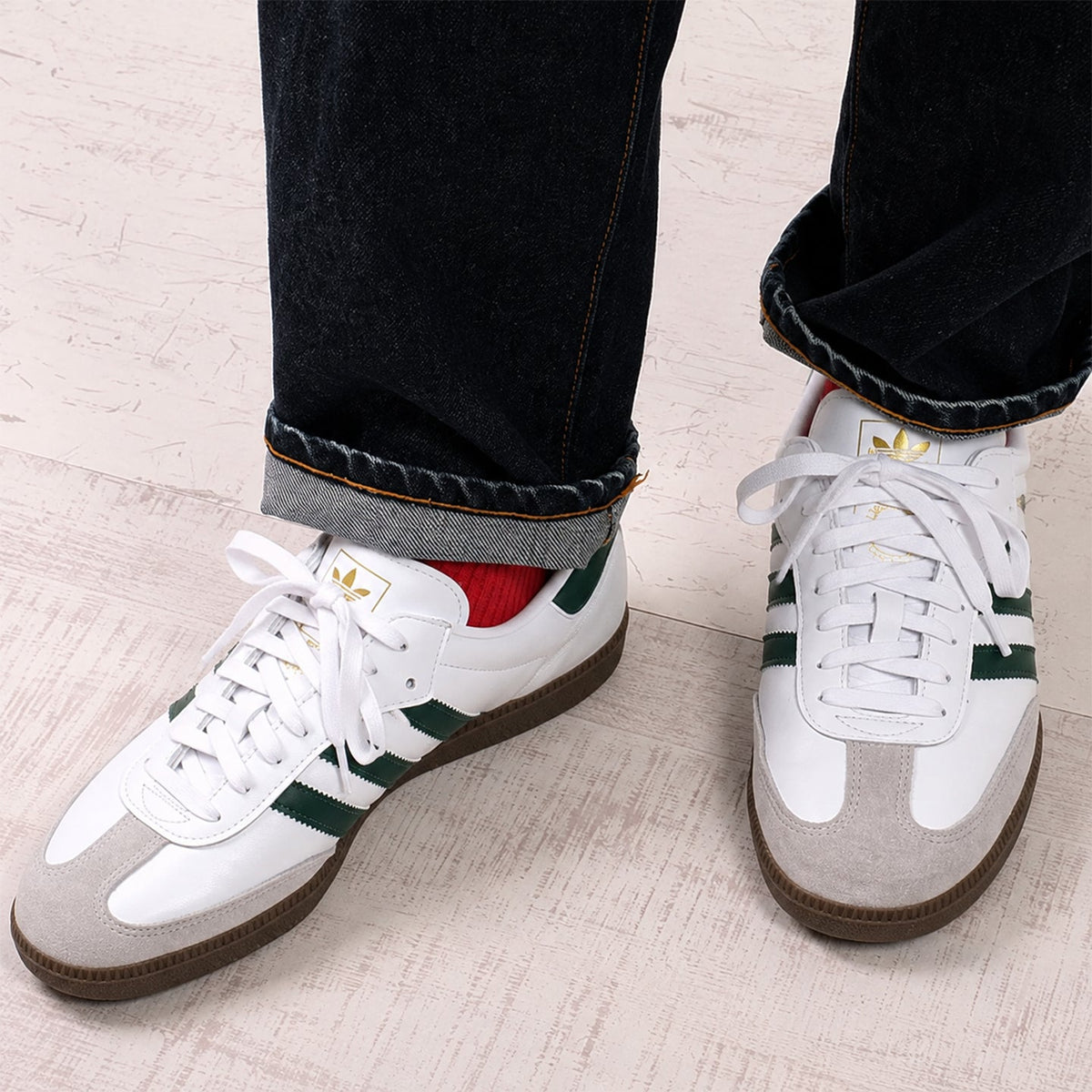 White Adidas Samba sneakers with green stripes and a light gray suede toe cap, shown on a person wearing dark cuffed jeans and a red sock, standing on a light wood floor.