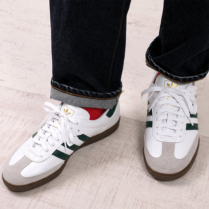 White Adidas Samba sneakers with green stripes and a light gray suede toe cap, shown on a person wearing dark cuffed jeans and a red sock, standing on a light wood floor.