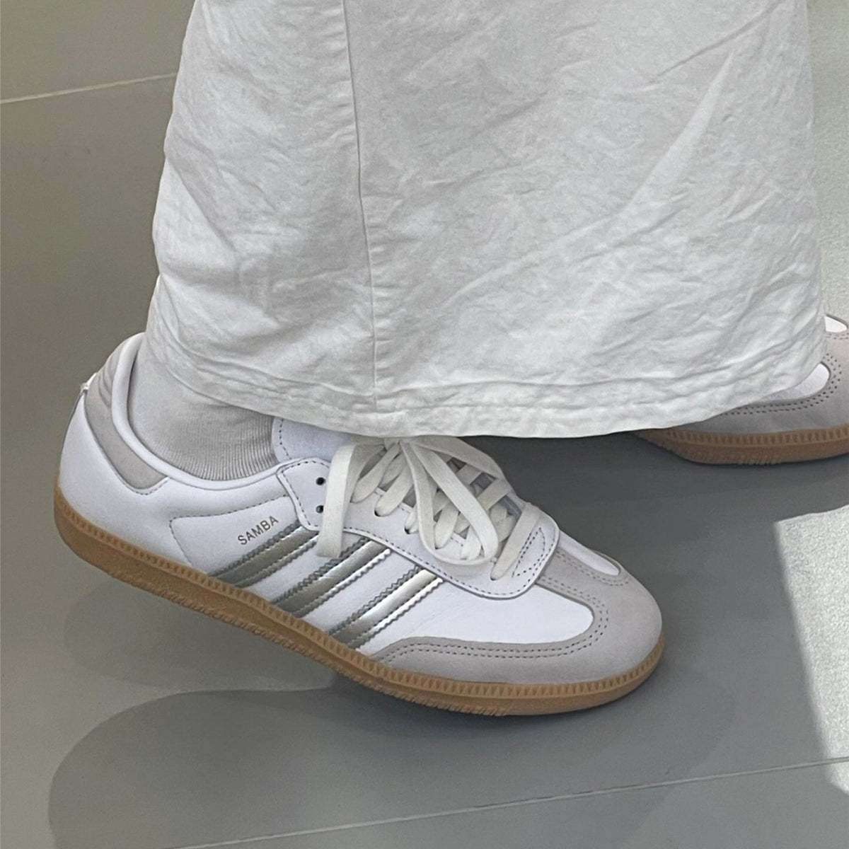 White Adidas Samba sneakers with silver stripes and gum soles, shown on a person wearing white pants.