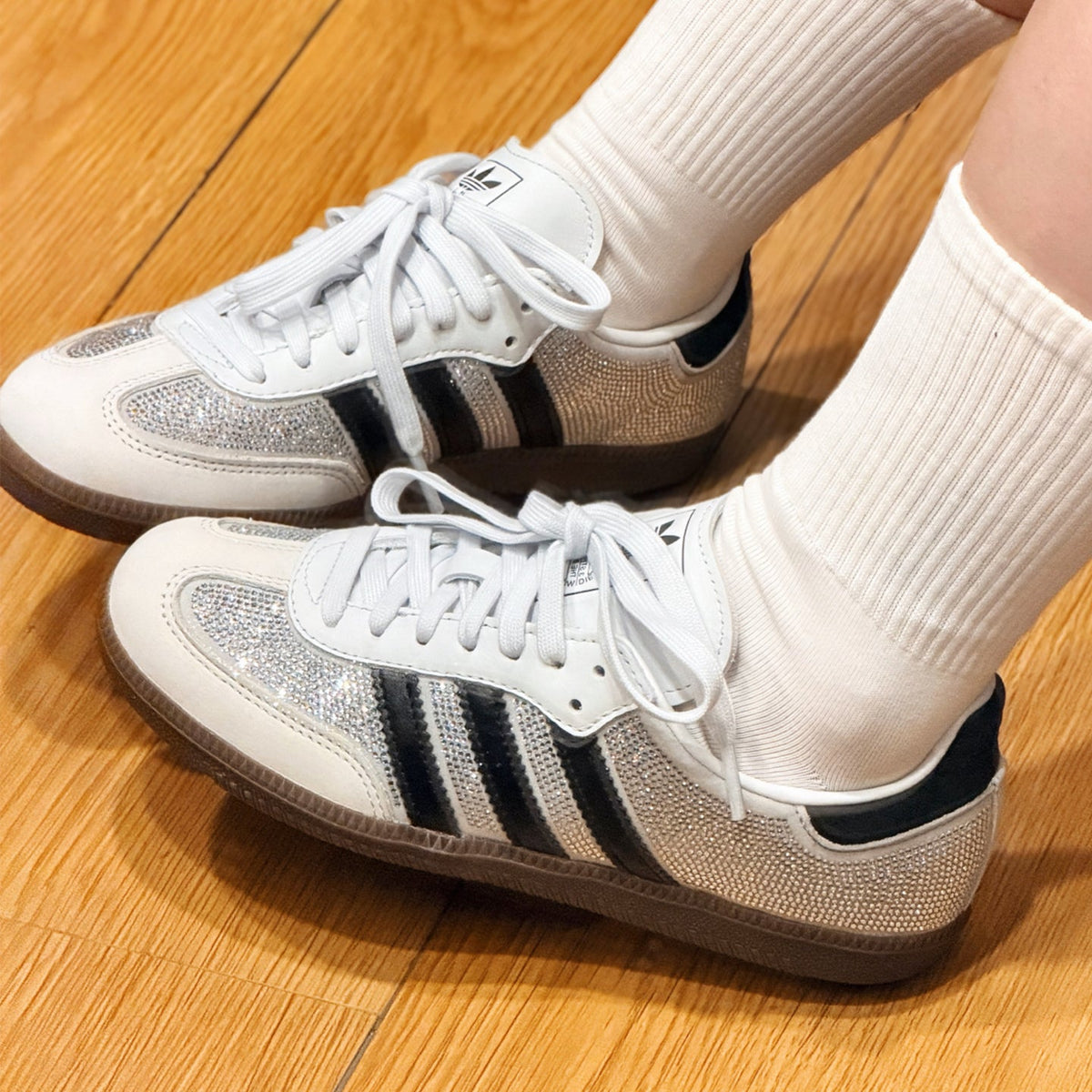 White Adidas Samba sneakers with silver glitter panels and black three-stripes, worn with white crew socks on a wooden floor.