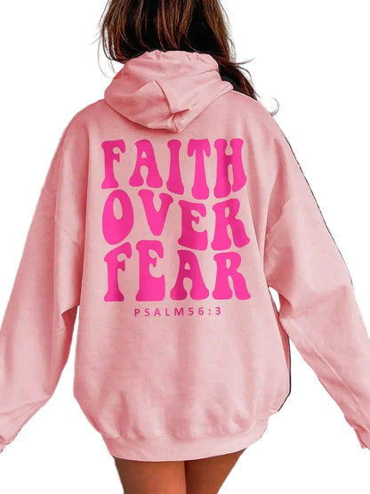 A person shown from the back wearing an oversized light pink hoodie with large bright pink text reading 'FAITH OVER FEAR' and smaller 'PSALM 56:3' printed on the back.