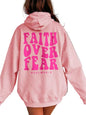 A person shown from the back wearing an oversized light pink hoodie with large bright pink text reading 'FAITH OVER FEAR' and smaller 'PSALM 56:3' printed on the back.