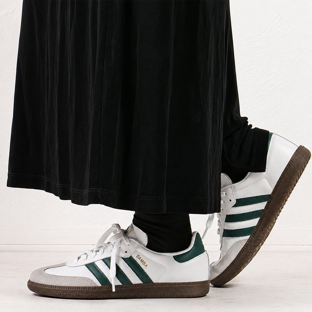 White Adidas Samba sneakers with dark green stripes and heel tab, worn with black socks and a black pleated skirt against a white studio background.