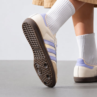 A close-up of a person wearing cream and light purple sneakers with white ribbed socks, shown from the side and sole on a clean studio floor.