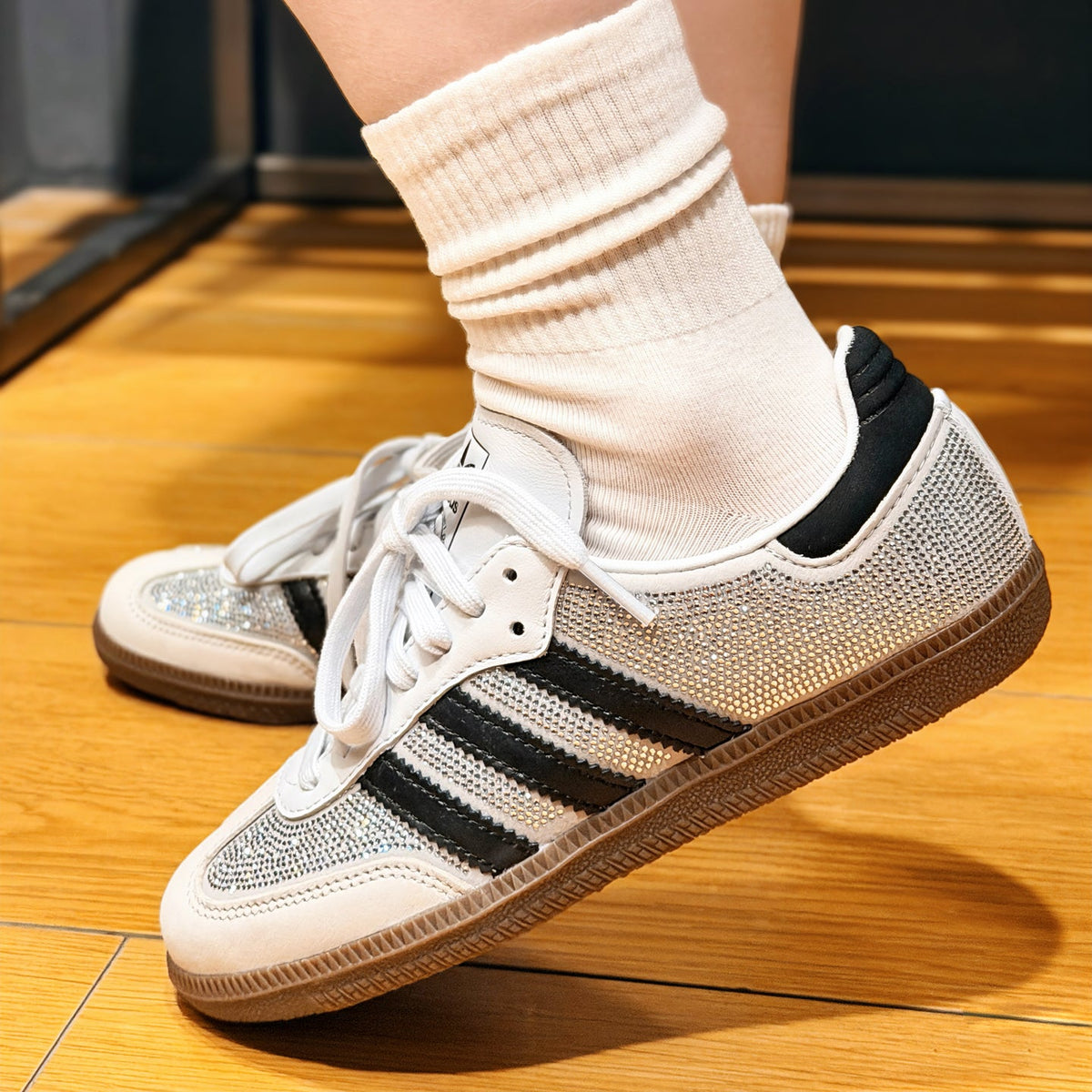 A person wearing white crew socks and white low-top sneakers with black side stripes and a textured knit upper, standing on a wooden floor.