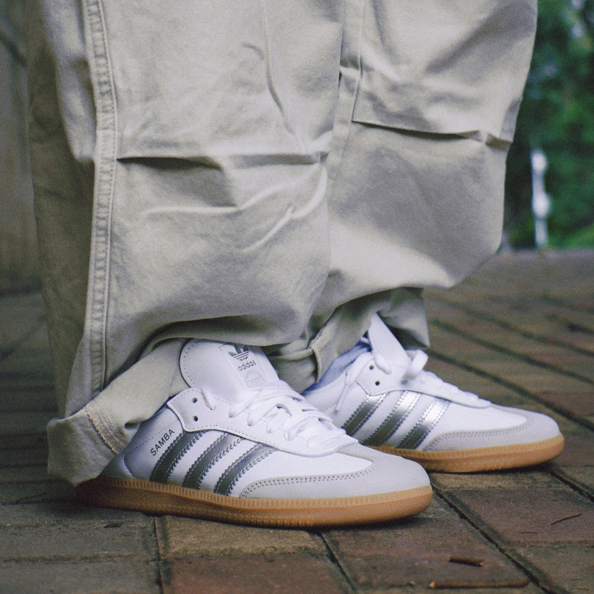 White Adidas Samba sneakers with metallic silver stripes and gum soles, worn with beige cargo pants on a brick patio.