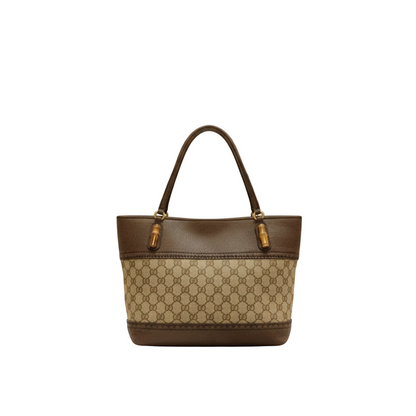 A structured tote handbag with a brown and beige monogram pattern, featuring dark brown leather trim and handles with gold-tone hardware, displayed on a white marble counter in a softly blurred boutique setting.