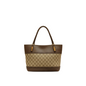 A structured tote handbag with a brown and beige monogram pattern, featuring dark brown leather trim and handles with gold-tone hardware, displayed on a white marble counter in a softly blurred boutique setting.