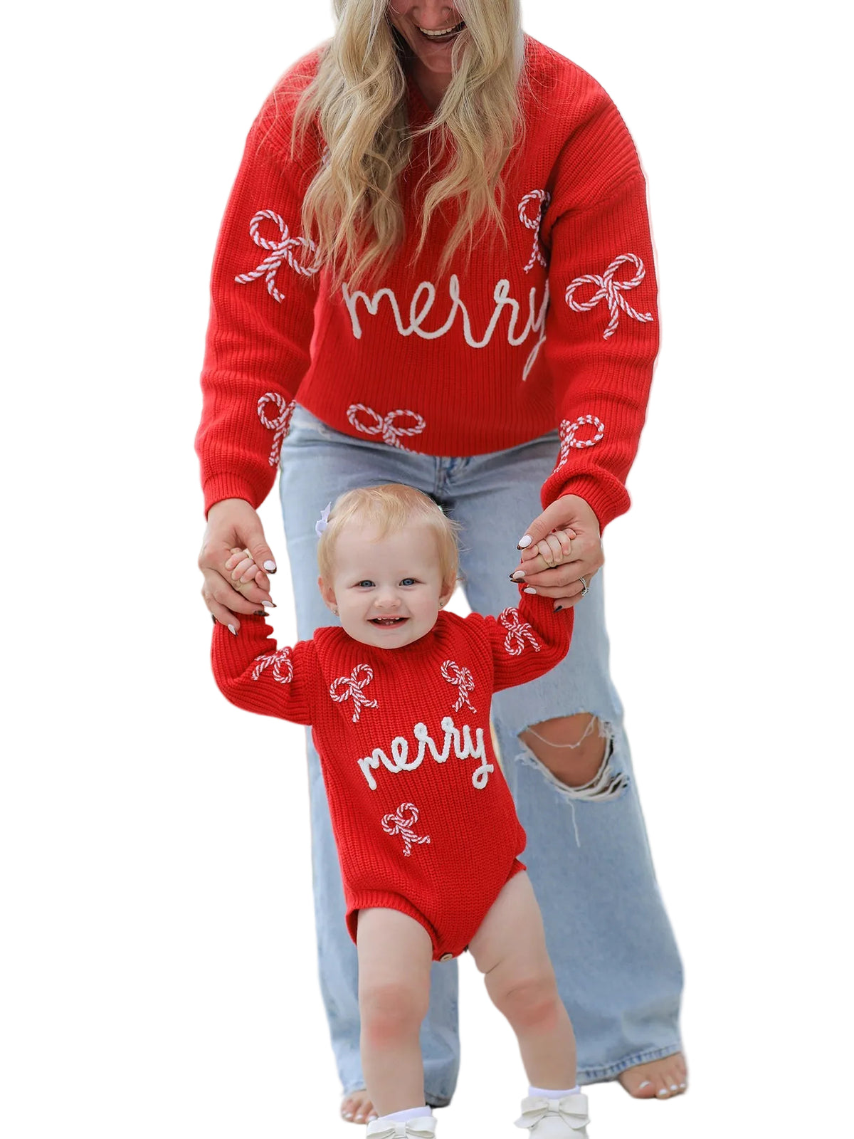 A woman and a baby wearing matching red knit sweaters with white 'merry' script and bow patterns, standing outdoors on a paved path with a blurred field and trees in the background.
