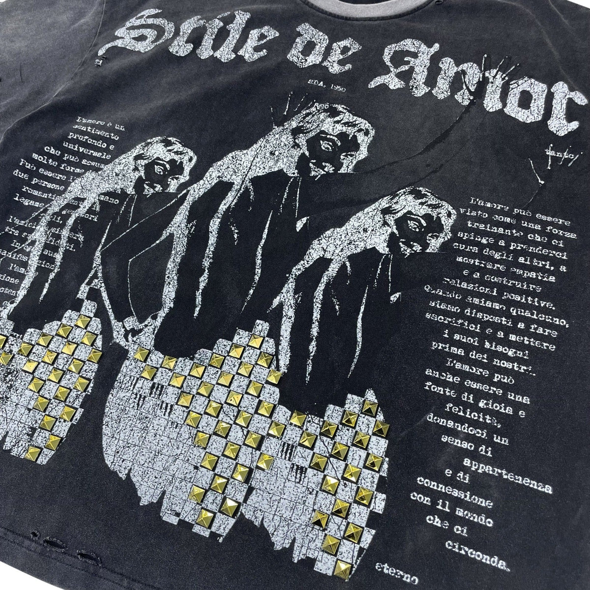 Close-up of a black graphic T-shirt featuring the band name 'Gole di Amor' in distressed white lettering, a stylized white-and-black illustration of three figures, Italian text, and a grid of gold-tone studs across the lower left.