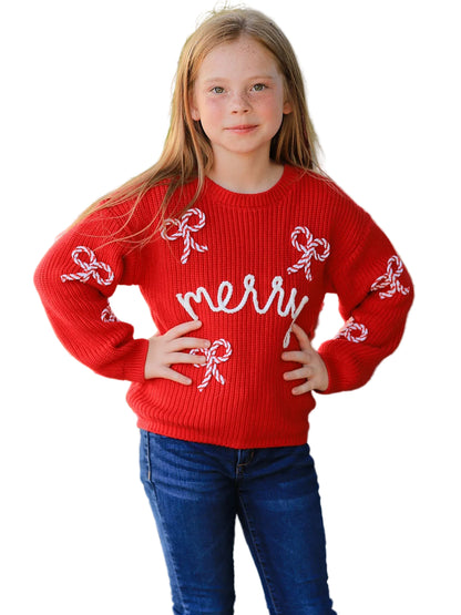 A young girl with long light brown hair stands outdoors wearing a bright red ribbed knit sweater with white embroidered bows and the word 'merry' across the chest, paired with blue jeans.