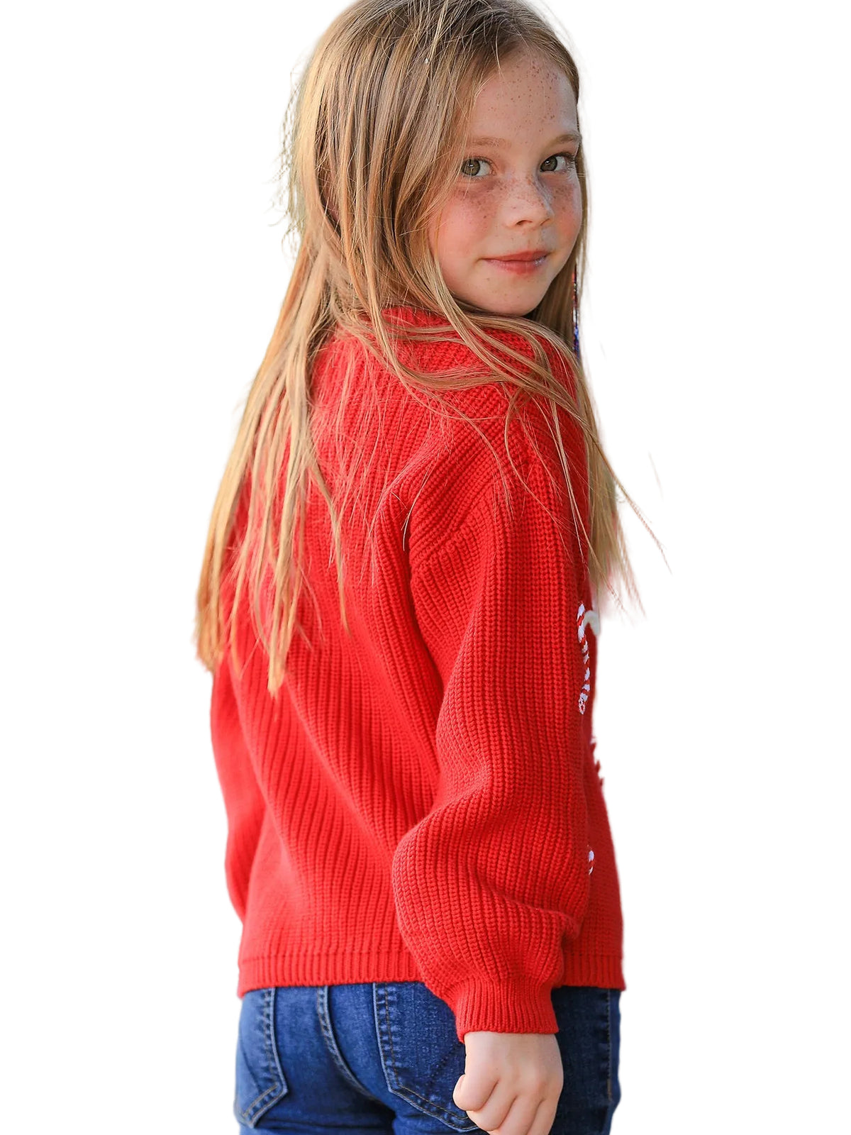 A young girl with long blonde hair is shown from the back wearing a bright red ribbed knit sweater and blue jeans, standing in front of a softly blurred outdoor background with red flowers.