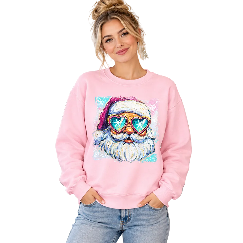 A woman wearing a light pink crewneck sweatshirt with a colorful graphic of a Santa Claus face wearing heart-shaped sunglasses, standing in a decorated holiday kitchen. The brand name 'ORNAMENTALLYYOU' is visible in the bottom right corner.